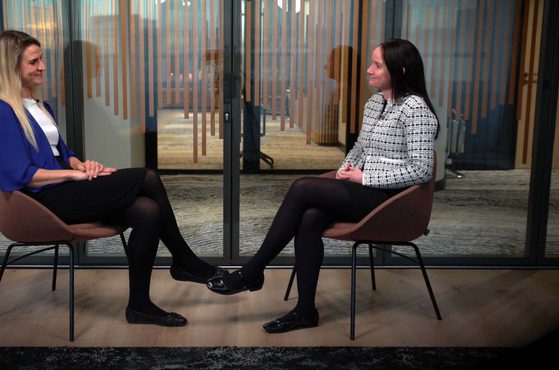 Two women sit facing each other in a modern indoor setting, separated by a glass wall with wooden design elements, engaging in conversation.