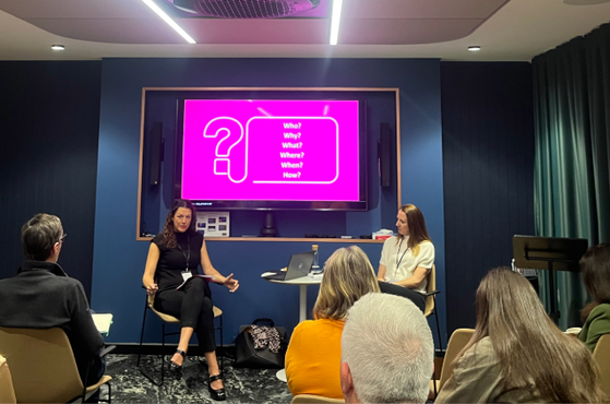Two panelists on a blue-walled stage face an audience. A vivid pink screen behind them shows a large question mark and questions: Who, Why, What, Where, When, How.
