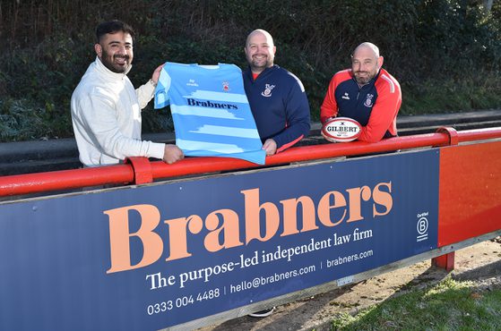Three men stand behind a sports barrier, smiling. One holds a blue sports jersey, another has a rugby ball, and they are outdoors with trees in the background.