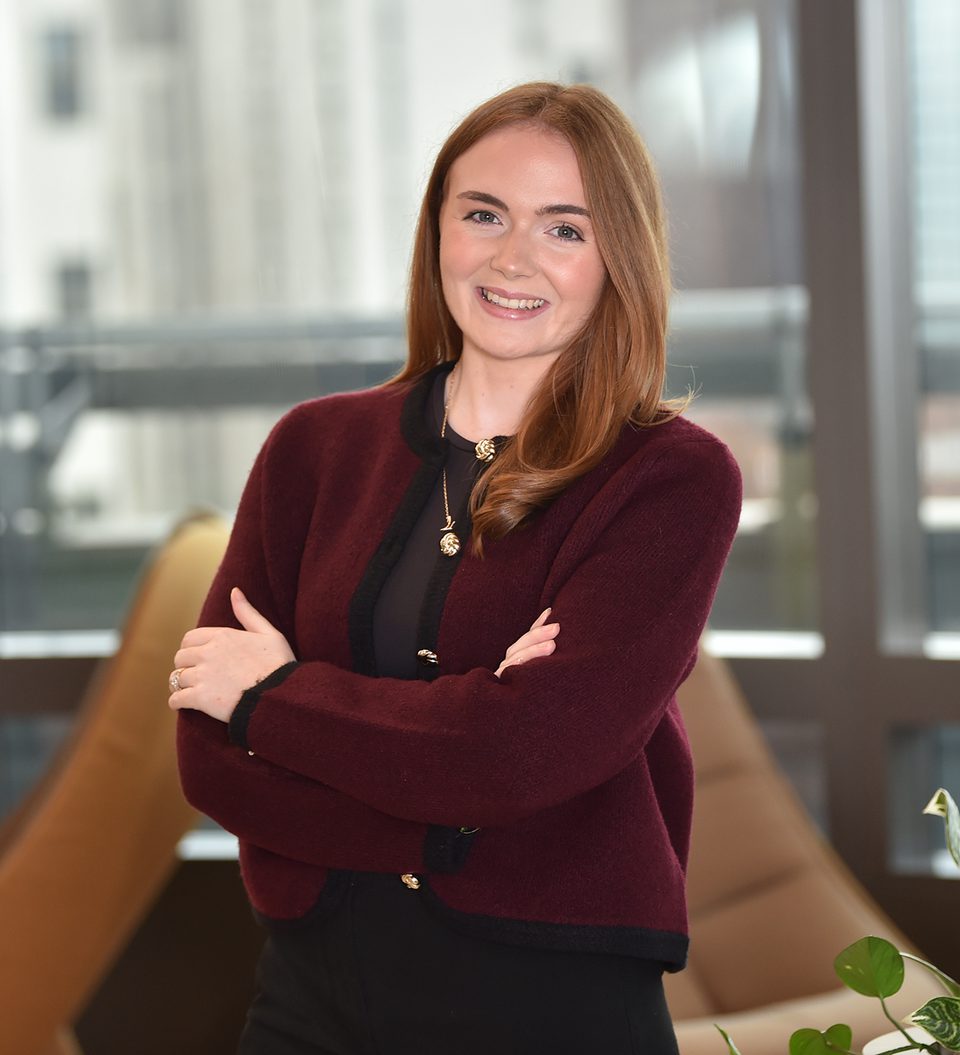 A woman with long red hair, smiling, standing with crossed arms in an office space with large windows, a brown lounge chair, and green plant leaves.