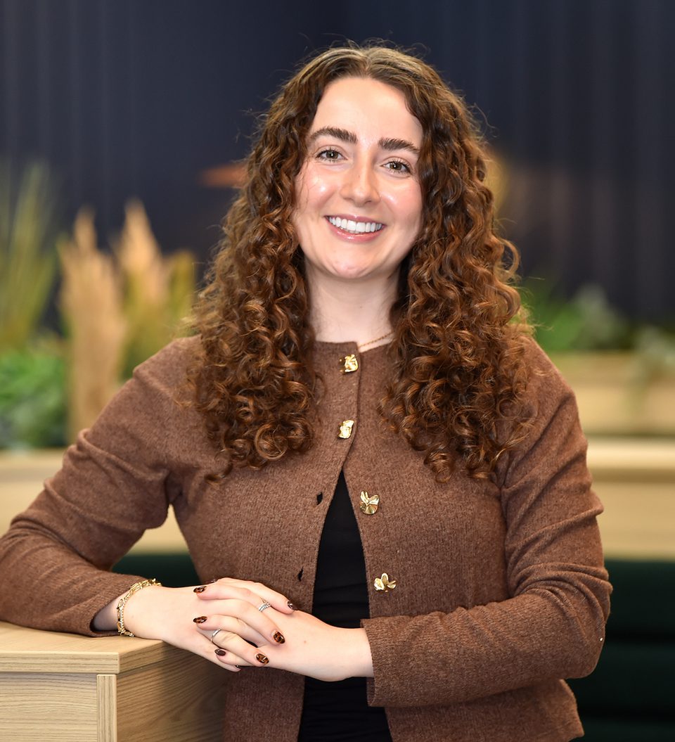A woman with long, curly brown hair, smiling and sitting at a wooden table in an indoor setting with blurred green plants and a dark background.
