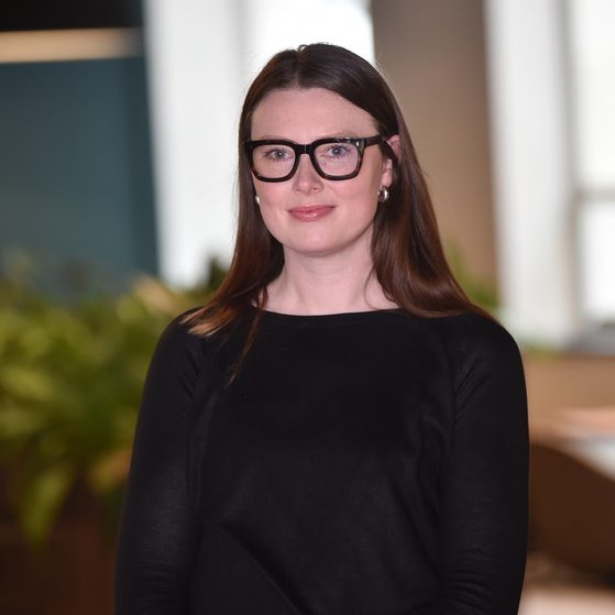 A woman with long brown hair, wearing black glasses, earrings, and a black top, standing indoors with a blurred background of a window and plants.