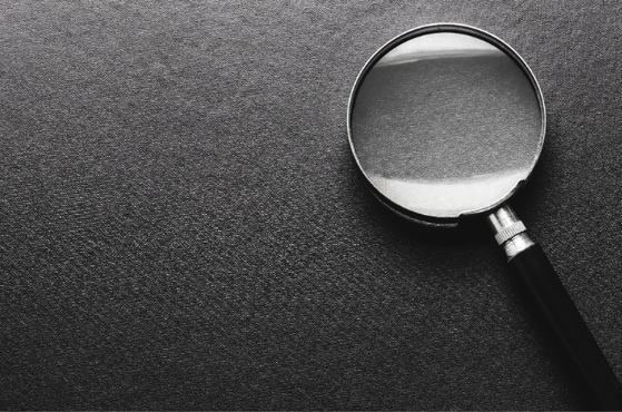 Black magnifying glass with a metal rim rests on a dark textured surface, angled toward the upper right; greyscale close-up highlighting the glass and handle.