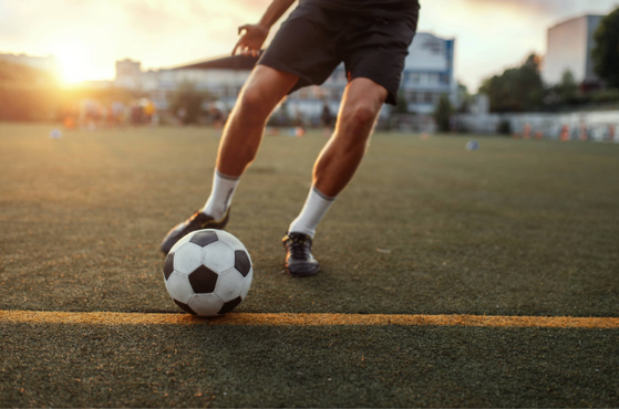 A person on a football field prepares to kick a black-and-white football at sunset, with grass surface and blurred background buildings.