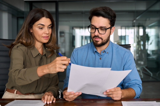 A woman and a man sit at a table discussing and reviewing documents in a modern office setting, with glass walls and an open-plan background.