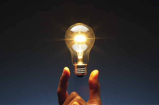 A lit incandescent light bulb held between the fingers of a hand, glowing against a dark blue background.