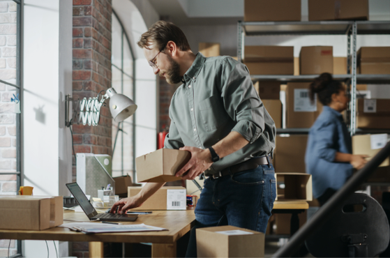 Person with glasses at a desk in a warehouse, typing on a laptop and packing a cardboard box, with shelves of boxes in the background.
