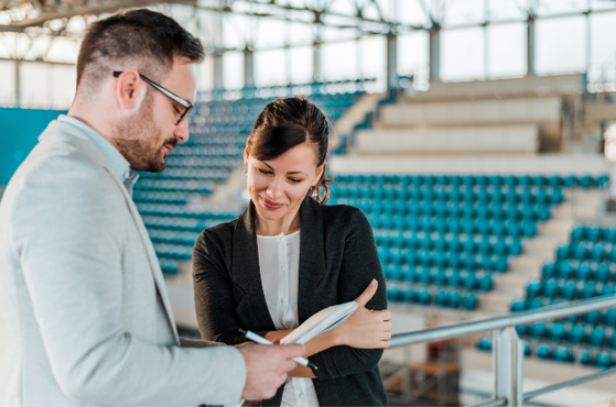 Two professionals in smart clothing review documents together in a modern indoor arena, standing by a railing with rows of blue seats behind them and a bright, airy ceiling.