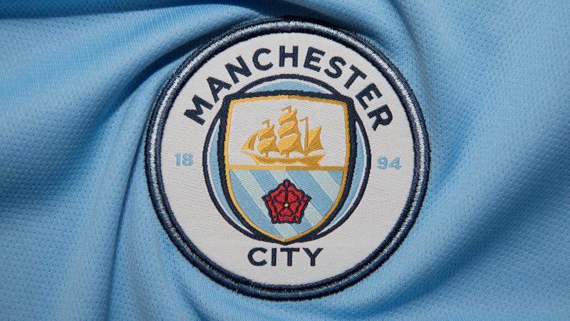 Close-up of a Manchester City crest badge on light blue fabric, featuring a gold sailing ship on a shield with a red rose and the words MANCHESTER CITY.