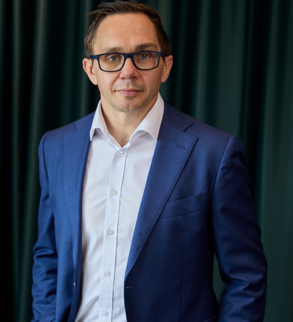 Person wearing a blue suit and white shirt with dark-framed glasses stands against a dark green curtain, looking at the camera with a neutral expression.