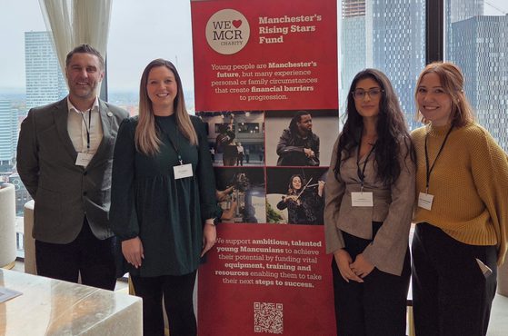Four people stand side by side in front of a red banner at a conference, with city buildings visible through large windows behind them.