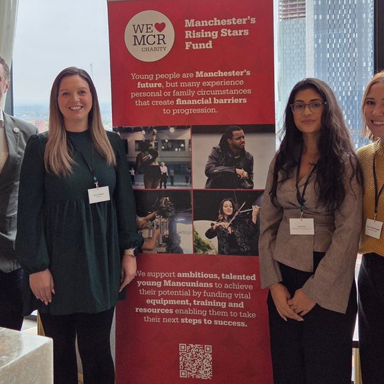 Four people stand side by side in front of a red banner at a conference, with city buildings visible through large windows behind them.