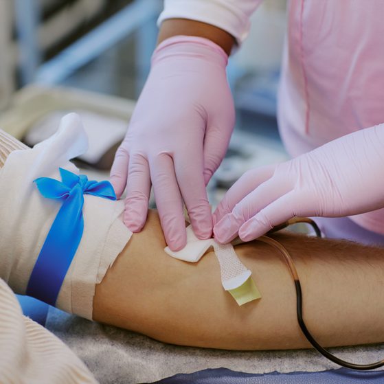 A healthcare professional in pink scrubs and pink gloves is inserting a needle into a patient's arm, which is secured with tape and a blue ribbon.