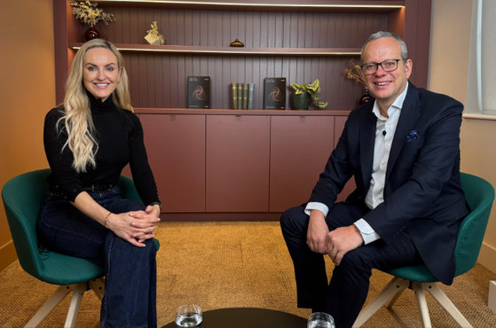 A woman and man sit on green chairs in an office, smiling at the camera with a small round table and glasses of water in front; background features shelves with decorative items.