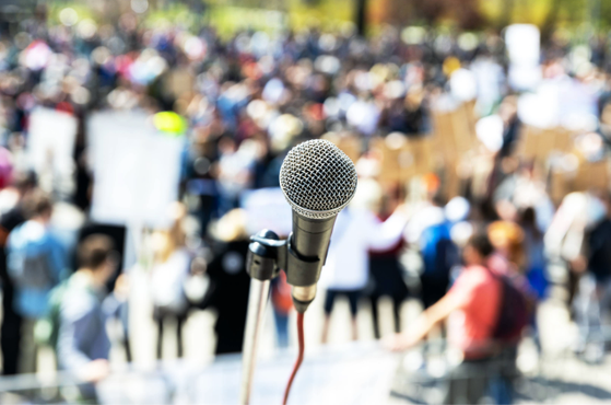 A microphone in the foreground with a large crowd of people holding signs and banners in the background during a protest or rally.