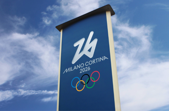 A tall blue sign featuring the Milano Cortina 2026 Winter Olympics logo, with the Olympic rings below, set against a partly cloudy sky.