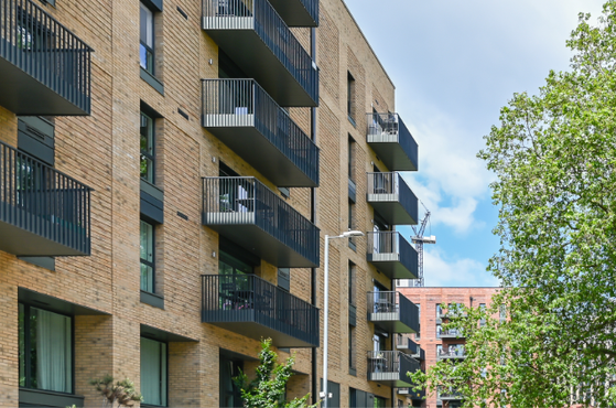 A multi-storey residential building with brick exterior and black balconies, surrounded by green trees and a partly cloudy blue sky.