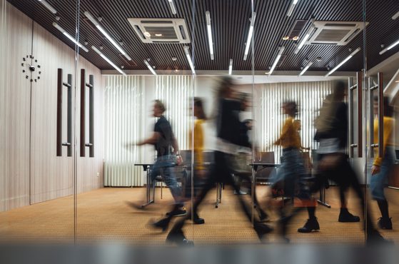 Blurred motion of people walking in a modern conference room with wooden wall panels, glass partition, black ceiling with linear lighting, and a clock on the wall.