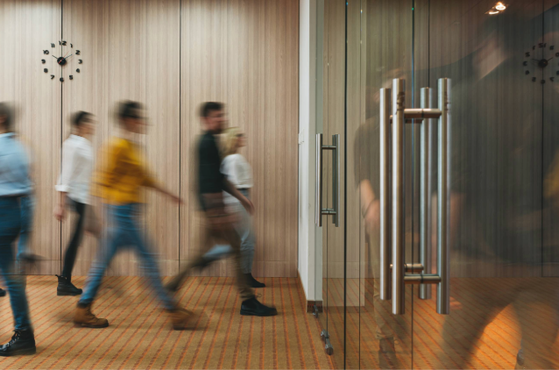 Blurred group of people walking in a line past a wooden-paneled wall with a clock, seen through glass doors, in an indoor setting.
