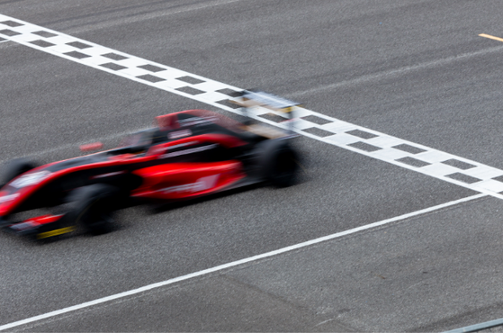 Blurry red Formula race car speeds along a grey tarmac track, crossing a black-and-white checkered finish line.