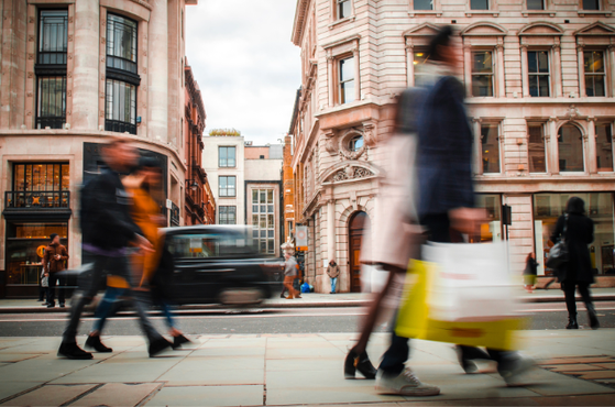 Blurred pedestrians walk along a busy city street in front of ornate beige stone buildings and shopfronts, while a moving vehicle passes by.