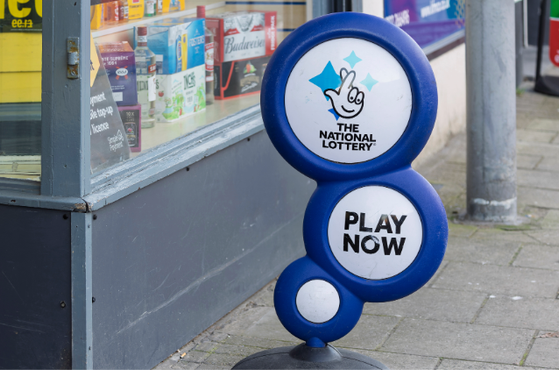 Two blue circular signs stacked for The National Lottery, featuring a white panel with a smiling hand logo and 'Play Now', mounted on a stand beside a shop window.