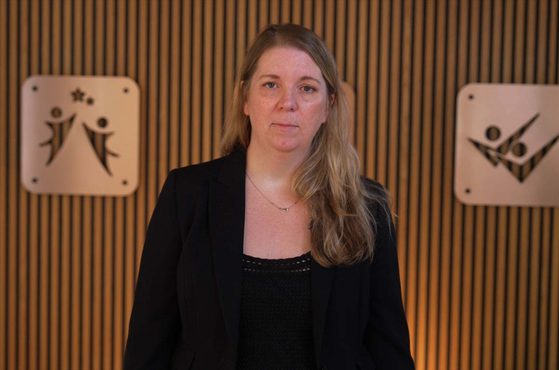 Person with long blonde hair wearing a black blazer and top stands in front of a vertical wooden slat wall, flanked by square signs with abstract human figures.