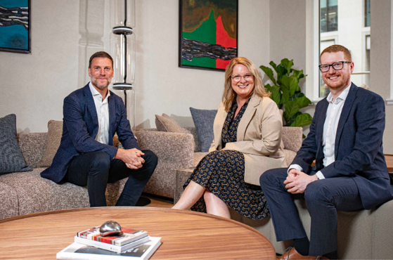 Three professionals sit on a grey sofa in a modern office lounge, smiling. A round wooden coffee table with magazines sits in front, with colorful abstract art on the walls.