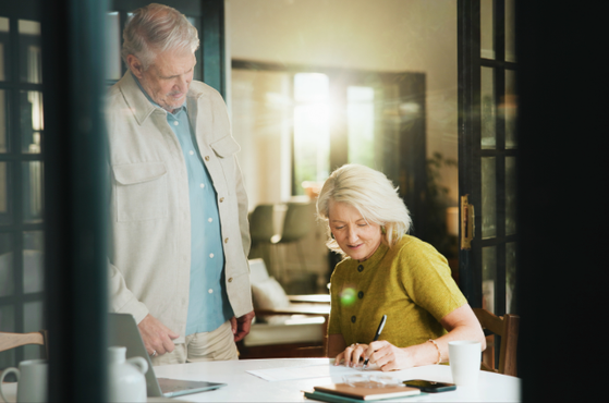 Two older adults in a bright, cozy room: one seated at a table writing in a notebook, the other stands nearby watching, with warm light streaming through glass doors.