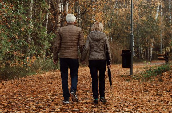 An elderly couple walking hand in hand along a leaf-covered path in a forest during autumn.