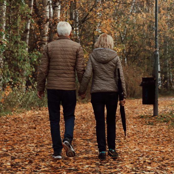 An elderly couple walking hand in hand along a leaf-covered path in a forest during autumn.