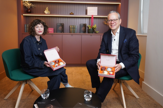 A woman and a man sitting in green chairs in a cosy office, each holding an open box of Christmas ornaments, with a small black table and glasses of water in front of them.