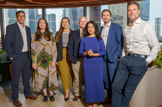 Seven diverse professionals in smart business attire stand together in a modern office, with a city skyline, large windows and greenery in the background.