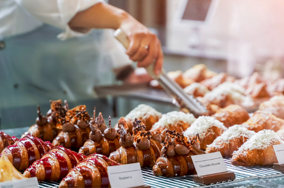 Display case of assorted bakery pastries under warm light, featuring chocolate-topped choux and glossy glazed rolls; a staff member uses tongs while flavour labels are visible.