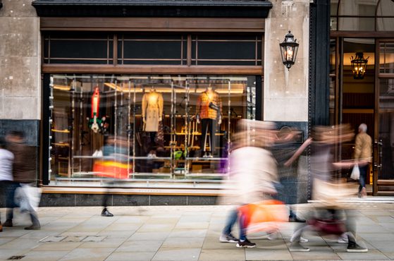 Blurred pedestrians walking past a shop window with mannequins displaying coats and accessories, set in a stone building with black accents and lantern-style lights.