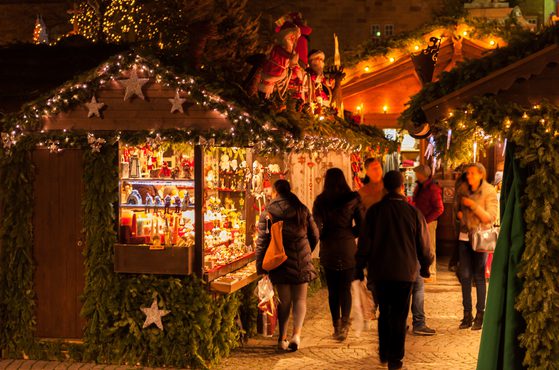 A cozy Christmas market at night with decorated wooden stalls, twinkling lights, and people browsing festive ornaments and gifts.