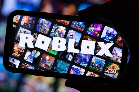 A hand holds a smartphone displaying the Roblox logo amid a grid of colourful game thumbnails on a vibrant, blurred background with small square Roblox icons filling the screen.