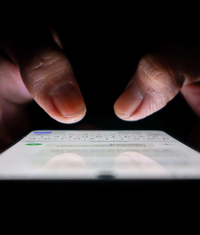 Close-up of hands tapping a glowing smartphone screen in a dark setting, with fingers hovering over the device as a messaging interface lights up.