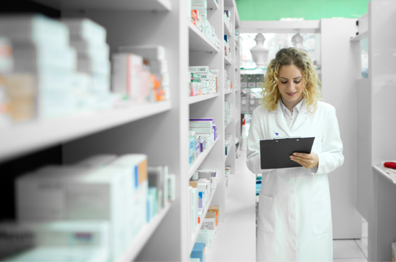 A healthcare professional in a white lab coat stands in a bright pharmacy aisle, holding a clipboard and reviewing documents among shelves of medicines.