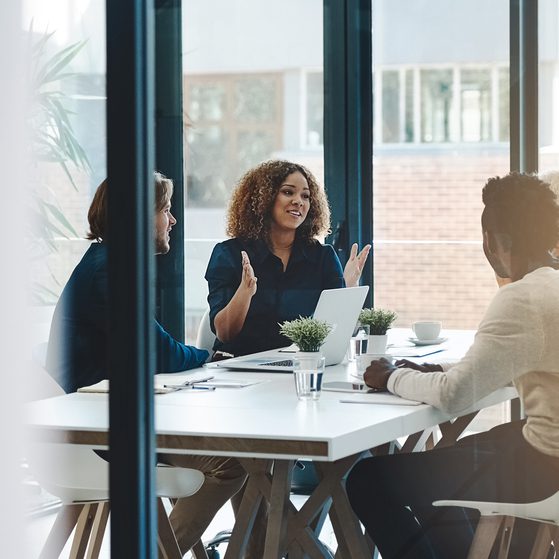 A woman with curly hair is speaking to a group of three colleagues in a modern office meeting room, with a whiteboard and large windows in the background.