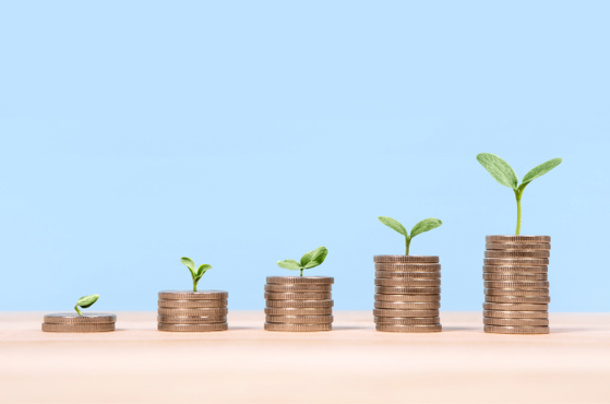 Series of coin stacks with small green sprouts growing taller from left to right on a wooden surface against a blue background.