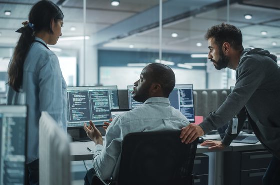 Three colleagues having a discussion in a modern office with multiple computer screens displaying code, with bright natural light and a glass wall in the background.