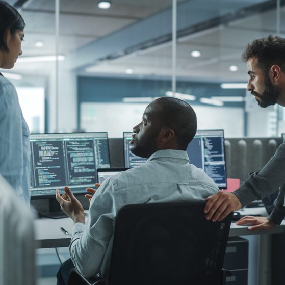 Three colleagues having a discussion in a modern office with multiple computer screens displaying code, with bright natural light and a glass wall in the background.