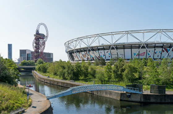 Clear blue sky over a canal with a blue pedestrian bridge, a curved stadium to the right, and the red ArcelorMittal Orbit sculpture in a green park.
