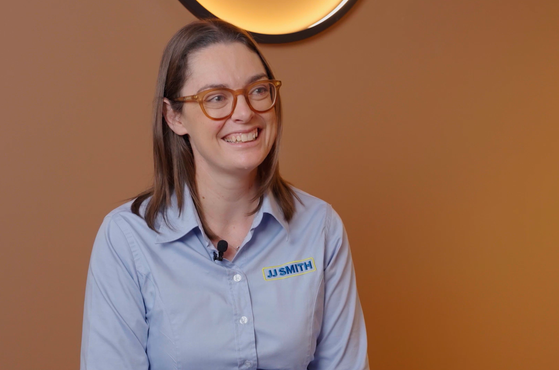 A woman with brown hair, glasses, and a light blue shirt with a name tag that reads "JJ SMITH" is smiling and sitting against an orange wall with a circular light fixture above her.