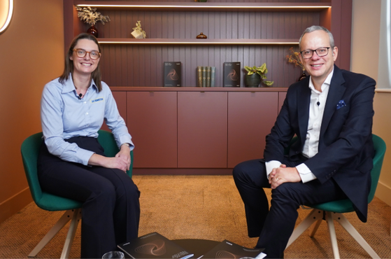 A woman and a man sit in modern chairs in a room with warm lighting and a bookshelf-style background, smiling at the camera during a professional meeting.