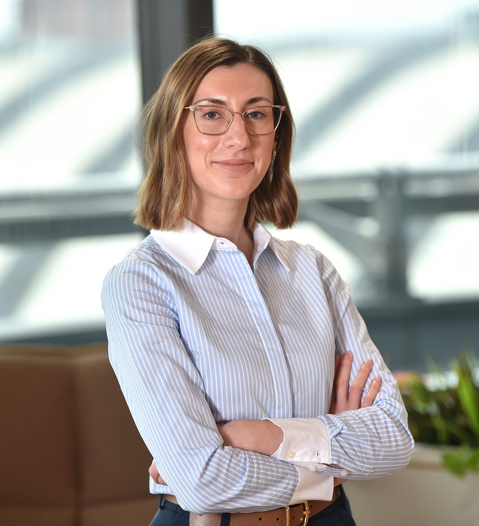A woman with shoulder-length brown hair, wearing glasses and a blue-and-white striped shirt, standing with arms crossed in a modern office.