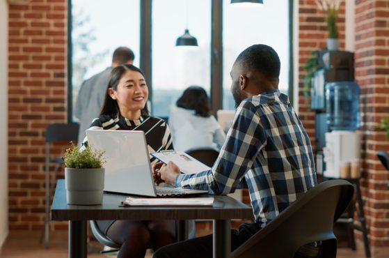 A man and a woman are seated across from each other at a desk in a modern office with brick walls, large windows, and plants, engaged in conversation.