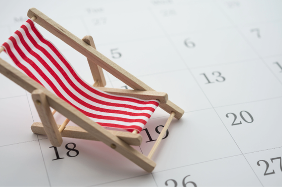 A small wooden beach chair with red and white striped fabric sits on a calendar page, suggesting summer holidays and planning for upcoming dates.