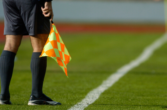 Assistant referee on a grassy football pitch near a white touchline, wearing black shorts and socks, holding an orange and yellow checkered flag.